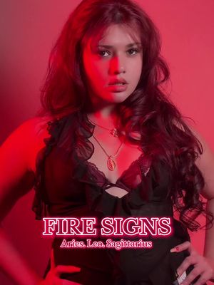 A woman is centered, framed from mid-torso up, against a red backdrop. She has long, wavy auburn hair and is wearing a black, lacy-trimmed top. A gold necklace with a pendant rests on her chest. The image features a neon-style text overlay: "FIRE SIGNS" with the zodiac signs Aries, Leo, and Sagittarius below. The woman is looking at the camera with a subtle expression.