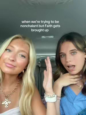 A selfie-style shot taken from inside a car features two young women. The woman on the left has blonde hair and is wearing a gold necklace with a cross pendant, with a necklace. The woman on the right has longer brown hair, wearing a necklace and has her mouth open. Text overlayed on the top reads 'when we're trying to be nonchalant but Faith gets brought up'.