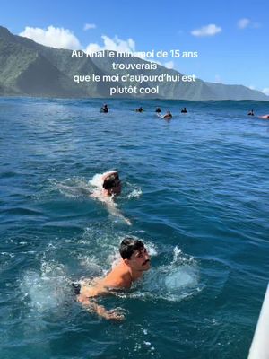 Two men are swimming in a clear blue ocean. The foreground features the men, with one closest to the camera sporting a mustache. The background shows a green mountain under a clear blue sky, and several other people floating in the water. Text overlays the image.