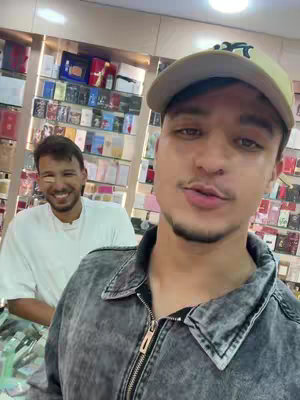 A selfie-style shot taken inside a store, showcasing two men. The main subject is a man facing the viewer, wearing a cap and a denim jacket. Behind him, another man smiles broadly. Shelves filled with products are visible in the blurred background.