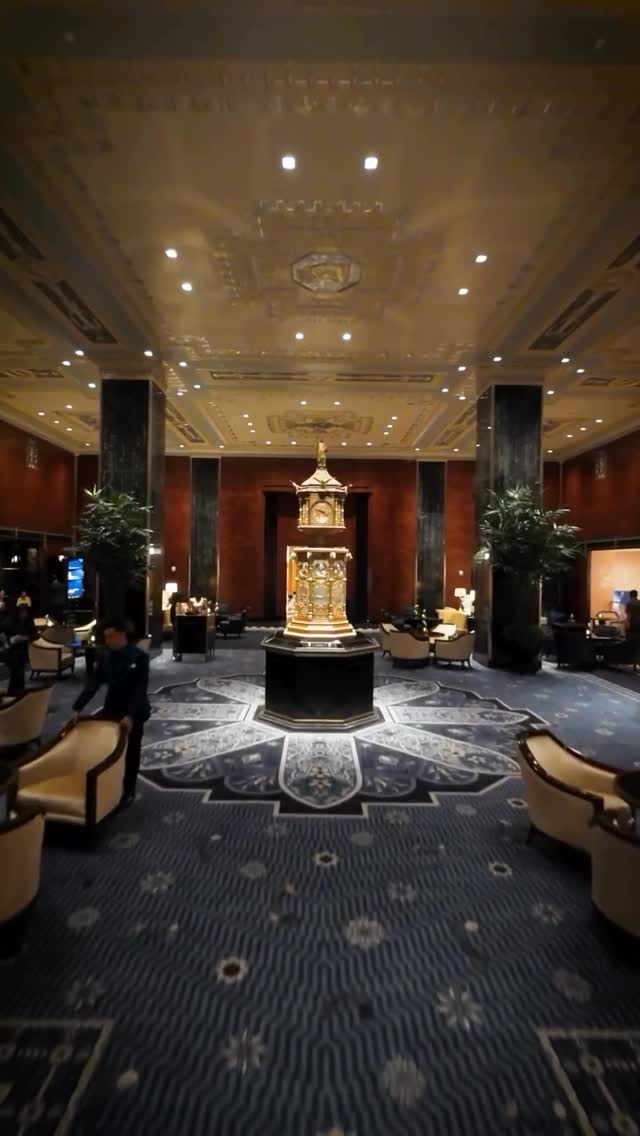 An interior shot of a grand hotel lobby. The focus is on a large, ornate clock that sits on a pedestal in the center. The floor has a patterned rug, and there are armchairs and plants visible. The color palette is warm, featuring gold, red, and dark blue.