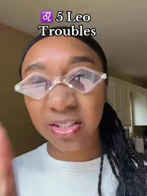 The cover image focuses on a Black woman's face from the chest up, looking slightly upwards. She wears a white t-shirt, clear-rimmed diamond-shaped glasses. Her makeup is visible, with glossy pink lips, and her black hair is styled with multiple braids pulled back. The text overlay at the top states "5 Leo Troubles" with a purple Leo zodiac symbol.