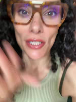 Close-up shot of a woman with curly dark hair and large glasses, waving at the camera. She's wearing a green tank top, and her mouth is slightly open, suggesting she's speaking. The focus is on her face and expression.