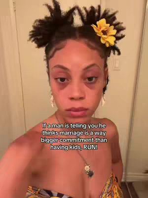A selfie-style shot of a woman with her hair styled in two buns, adorned with a yellow flower. She has noticeable eye makeup and is wearing a top. Text overlayed at the bottom reads, 'If a man is telling you he thinks marriage is a way bigger commitment than having kids, RUN!'