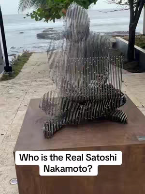 A metal sculpture of a seated figure on a pedestal. The sculpture is made of thin, vertical metal rods, giving a fragmented appearance. The figure is sitting with its legs crossed, apparently using a laptop. The sculpture is placed on a wooden pedestal. The background includes a beach, ocean, and a paved walkway.