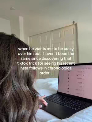 A woman with long brown hair is seen from the back and slightly from the side, sitting on a bed with a laptop in front of her. The laptop screen is open, displaying content.