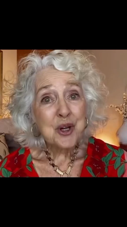 An elderly woman with short, wavy gray hair and fair skin looks directly at the camera, speaking with a slightly open mouth. She's wearing a red and green floral print top and a gold-colored chain necklace.