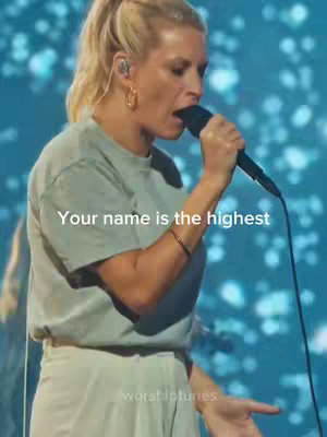 A woman with blonde hair and a bun is singing into a microphone, wearing a light-colored t-shirt. The background is a blurred, blue and white light display. Text overlay: "Your name is the highest".