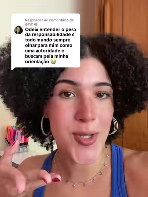 A young woman with curly black hair and hoop earrings is centered in the frame, looking directly at the camera. She wears a blue tank top and a delicate necklace, with a partially visible background that includes a desk and some colorful markers. A speech bubble with white text is superimposed on the image, and the creator is using a hand gesture.