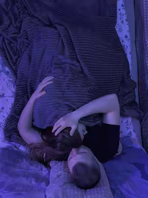 A top-down shot captures a close-up of two individuals cuddling in bed. The frame is bathed in a cool blue light. A crumpled gray blanket covers most of the scene. The person on top is obscured by the covers, with only their hand and hair visible. The other person's face is clearly visible beneath the covers. The color scheme is dominated by blues and grays.