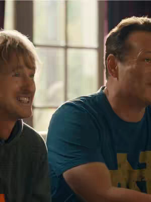 The cover image shows two men, presumably friends, in a living room setting. One is on the left, with short blond hair and a light green sweatshirt. He's smiling, showing his teeth, and looking to his right. The other man, on the right, has short, dark hair and is wearing a blue t-shirt. Both men are in a casual, relaxed pose indicating they're enjoying themselves.