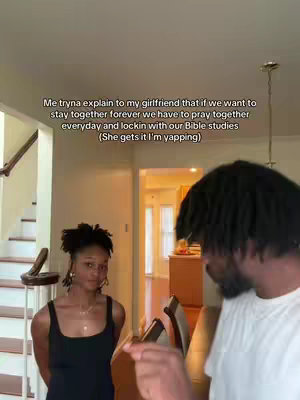 A man in a white t-shirt and dreadlocks is speaking in the foreground while his girlfriend, wearing a black top, stands looking at him, with a room and stairs in the background.