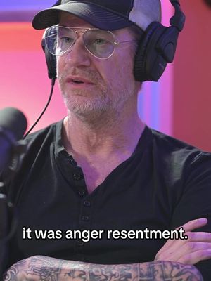 A man is seated in a recording studio setting, wearing glasses, a baseball cap, and headphones. The background is brightly lit with shades of pink and red. The overlaid text in white reads, "it was anger resentment."