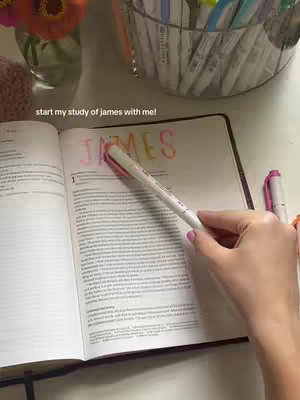 The image shows an open Bible on a light-colored surface, displaying the text "JAMES" highlighted in pink and orange over the first chapter. A hand, with visible fingernail polish, holds a white pen with a pink cap over the text. In the background, there is a clear glass container holding various colorful pens and highlighters, along with flowers in a vase.
