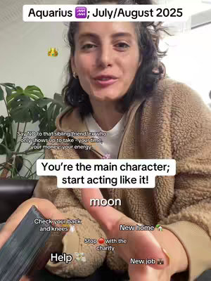 A woman is filmed from the waist up, centered, directly facing the camera, speaking. The background is blurred, but suggests an indoor setting with a plant visible, and what appears to be a dark colored couch. A large amount of text has been overlaid onto the video.  She is wearing a casual, light-colored, fleece-lined jacket. Her dark brown curly hair is neatly styled, and she's wearing a small gold hoop earring in her left ear. She is holding what looks like a Tarot deck on her left hand.