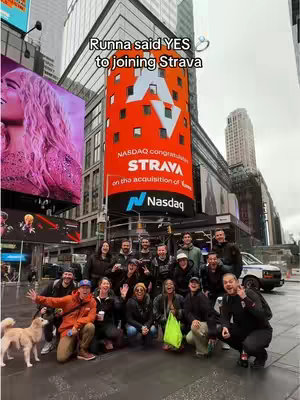 A group of people, likely a team, posing with a dog for a photo on a city street, under a large billboard. The billboard displays the Nasdaq logo and mentions Strava and Runna. A large building with glass windows is behind them.