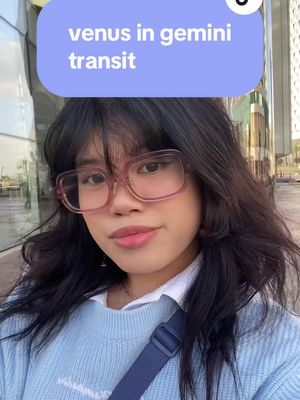 The cover image is a selfie featuring a young woman with dark hair and glasses. She is positioned in front of a building with tall windows, suggesting an urban setting. She has light skin, and her hair is styled with a layered cut. She is wearing a light blue sweater, a white collared shirt is slightly visible underneath. Above her head is a text overlay in a rounded rectangle that says, "venus in gemini transit."