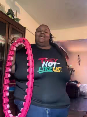 A woman stands indoors holding a pink, segmented hoop. She's wearing a black t-shirt with white and colored text, blue jeans, and her hair is styled in long braids. The background shows a living room setting with furniture and decor.