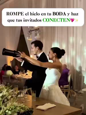 A photograph of a couple at their wedding reception. The bride and groom stand, the bride in a white strapless wedding dress and the groom in a black tuxedo and bow tie. The couple is holding a black object resembling a telescope, with the groom positioning it in front of the couple, as if they are collectively using it. They are clearly in a festive mood, and there are guests sitting at tables in the background. Uplighting creates soft glowing effects in the background.