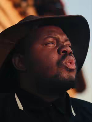 A medium shot features a man wearing a dark hat and a black shirt, with a neutral background. He has dark skin and a short beard with his lips pursed as if he is blowing a kiss. Above him is the edge of a building with what seems like a beige wall.