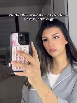 A woman holds a phone up to take a selfie in front of a mirror. The phone case has pink and white details. She is wearing a light gray jacket over a white top and has medium-length dark hair. The background includes a glimpse of a bedroom with white walls and a possible bed frame.
