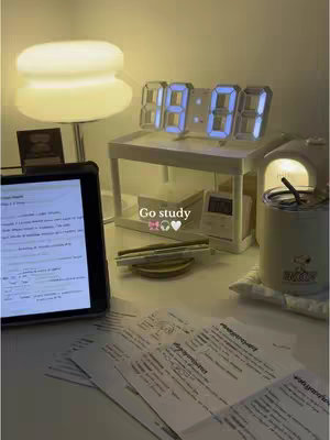 A well-lit desk setup for studying. There is a white desk with notebooks and papers scattered across the surface. In the background is a digital clock on a stand, and a lamp.