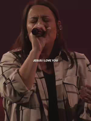 A woman with dark hair is singing into a microphone. The background is dark and blurry, with a red-brown color.