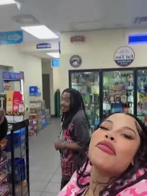 A selfie-style video taken in a convenience store setting. A woman is in the foreground with her face taking up most of the frame, smiling with full makeup. In the background, a man stands near shelves stocked with products. The overall composition suggests a casual, candid moment.