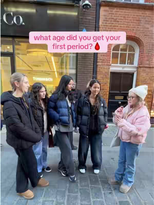 A group of young women are standing on a city street. A pink-colored overlay text bubble is near center, asking "what age did you get your first period?" The women appear to be wearing coats and casual attire. Brick building and storefronts form the background.