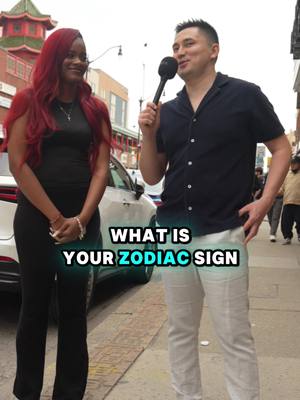 A man with a microphone interviewing a woman on a street. The woman has long red hair and is standing beside the man. Text overlay: "WHAT IS YOUR ZODIAC SIGN". They are in a city street, with a car in the background.