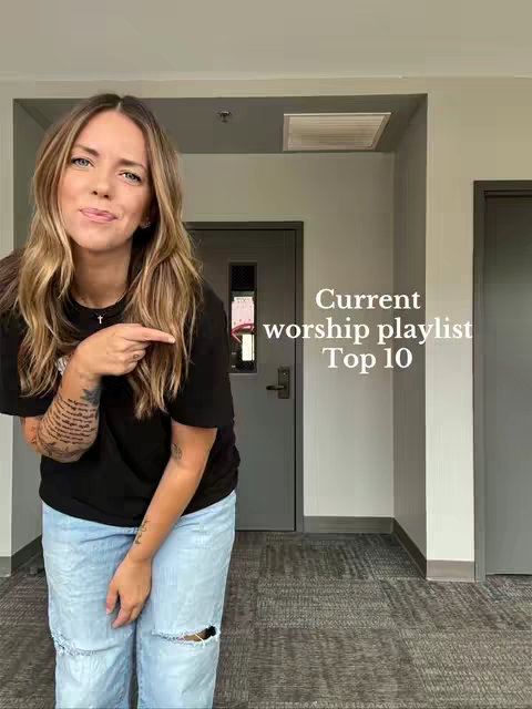 A woman with long brown hair stands in front of a gray door and wall, pointing to the right. She is wearing a black t-shirt and ripped jeans. The background is a simple indoor setting with neutral colors.