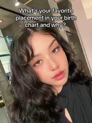 A selfie-style image featuring a young woman with dark, wavy hair and a light complexion. She has visible brow and lip makeup and a small nose ring. The background is blurred, which includes a sign on the left and the right, the walls are a beige-ish color, and the overall lighting is soft and natural.