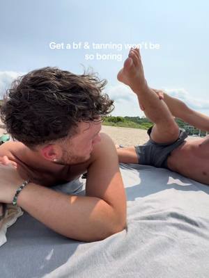 Two young men are lying on a beach towel on the sand, basking in the sun under a bright sky. The focus is on the one on the left, who is looking towards the other person. The other person is on the right, who has one leg up in the air. The image setting indicates a relaxed, sunny day.