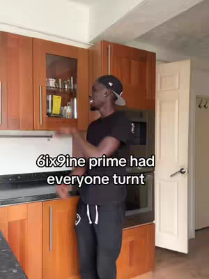 A person stands in a kitchen, likely filming a video. The person is wearing a black tee-shirt, black pants, and a black cap. Text overlaid onto the image states, "6ix9ine prime had everyone turnt". The kitchen has wooden cabinets and a stainless steel counter in the background.