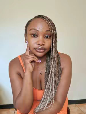 A young woman with long braided hair, wearing an orange top, is looking towards the camera. She is touching her face with a thoughtful expression. The background is a solid, neutral color.