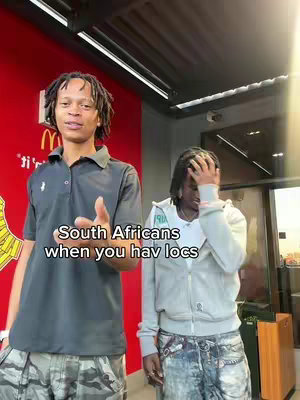 Two young men stand in front of a red background with a McDonalds sign. One man is on the left facing the camera with locs, and the other is on the right with his hand on his head. Overlaid text in white at the bottom says, 'South Africans when you hav locs.'