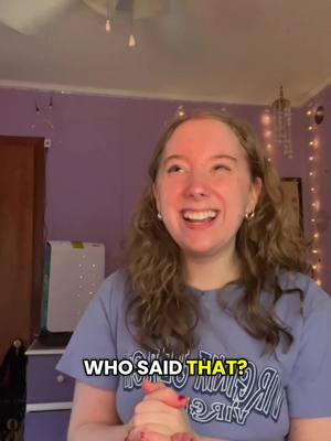 A woman with long wavy hair expressing joy, is smiling widely with her hands clasped. She's wearing a blue t-shirt with text on it. The background is a room with purple wallpaper, a wooden door, and a few decorative lights.