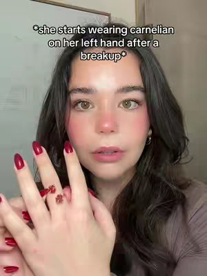 A woman with long dark hair and red nail polish is holding her left hand up towards the camera, showing off a ring with deep red gemstones. Text overlayed on the background reads, "she starts wearing carnelian on her left hand after a breakup."