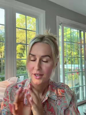 A woman with blonde hair in a half-up style is in a room with large windows. Natural light floods the space. She is wearing a patterned top, and her hands are clasped near her face. The background reveals a view of trees and sky.