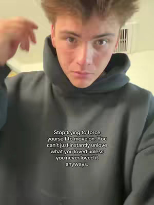 A young man with light skin, wearing a gray hoodie, is the central focus. The composition is a close-up shot, emphasizing his face and the upper part of his body. He is looking directly at the camera, with a neutral expression. The background is out of focus, suggesting an interior setting, with a hint of off-white wall. The text overlay is white and contrasts against the dark hoodie.