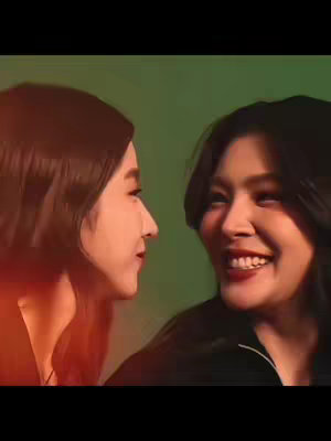 Two women are positioned closely, face-to-face with a green gradient background. The woman on the right has a large smile, indicating happiness, while the other looks toward her. A warm-toned filter is used, with a hint of red on the left side.