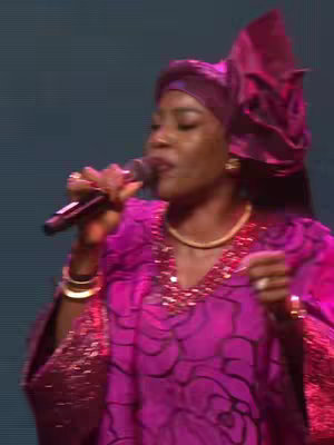 The cover image showcases a woman wearing a vibrant magenta dress and headscarf, speaking into a microphone. Her gold jewelry, including a necklace, earrings, and bracelets, adds to her appearance. The background is blurred, focusing attention on her, while she holds the microphone in her right hand.