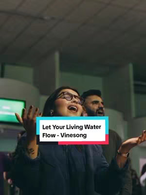 A vertical video cover features a woman in glasses and a man, both with their arms raised, appearing to sing or worship. A text overlay highlights the song title and artist, with a blurred background suggesting an indoor venue.