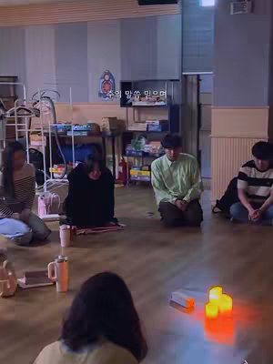 A group of people are kneeling on a wooden floor in a room, with some candles in the foreground. The background shows a room with shelves and a blue wall.