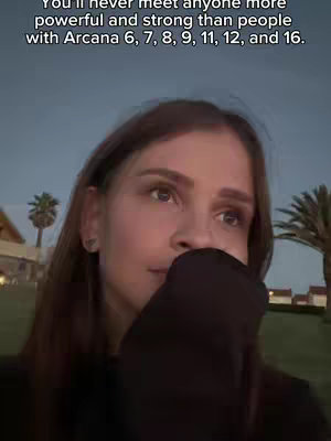 A medium shot of a woman with long, brown hair, looking down and to the side with a hand partially covering her mouth. The background is a blurry outdoor scene with trees and buildings, suggesting an evening or twilight setting. The woman is wearing a dark-colored hoodie, and the overall tone of the image is pensive and slightly melancholic.
