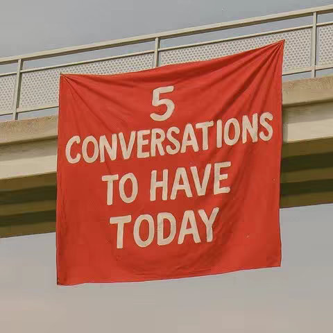 A red banner with white block letters hangs under a metal and concrete structure. The banner reads '5 CONVERSATIONS TO HAVE TODAY'. The sky is visible in the background.