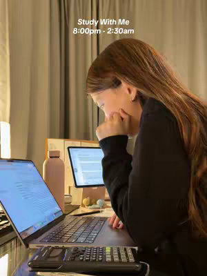 A young woman is seated at a desk, leaning towards a laptop. In front of her is a keyboard and a small calculator. A tablet with a document on it and a water bottle are also on the desk. Soft light is coming from behind the woman, and from a small desk lamp. The text overlay at the top of the image reads "Study With Me 8:00 pm - 2:30 am".