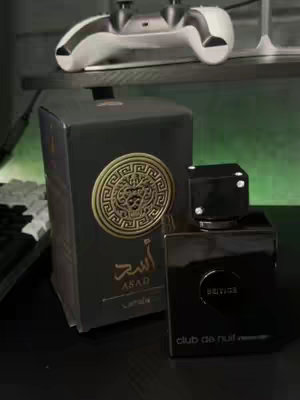 A dark-toned photo of perfume bottles and boxes. Two perfume bottles are on display, one with a black square cap and a label that reads "club de nuit." Beside those on the left side sits a black box for "Asad" brand perfume and a silver game controller sits on top of the box.
