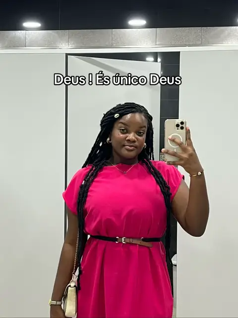 A selfie-style image taken in a bathroom. A black woman with long black braids stands facing the camera, holding a phone to take the photo, and wearing a vibrant pink dress with a black belt. The bathroom has a white and grey color scheme with a large mirror.