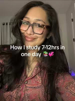 A selfie-style video cover image shows a person indoors. The person is a woman with long, dark hair wearing large-framed glasses that sit on a smiling face. She wears a lightly patterned, red-toned button-up shirt. The text overlay reads, 'How I study 7-12hrs in one day' over a book and heart emoji.